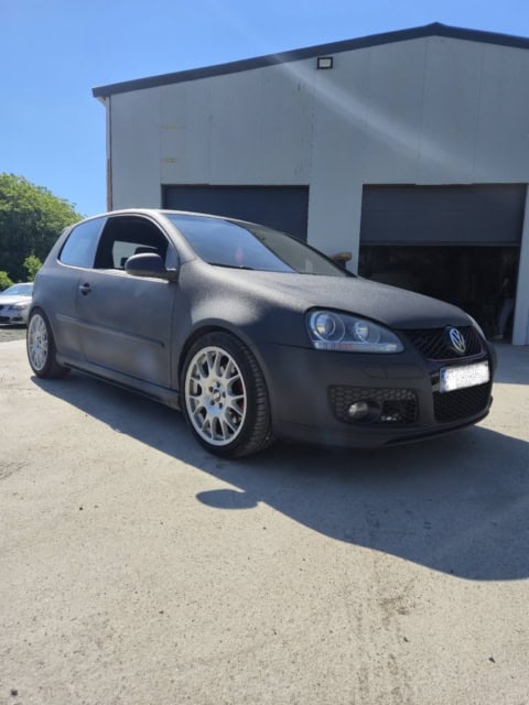 Mașină Volkswagen Golf neagră, sportivă, în fața unei clădiri industriale, pe o zi însorită.